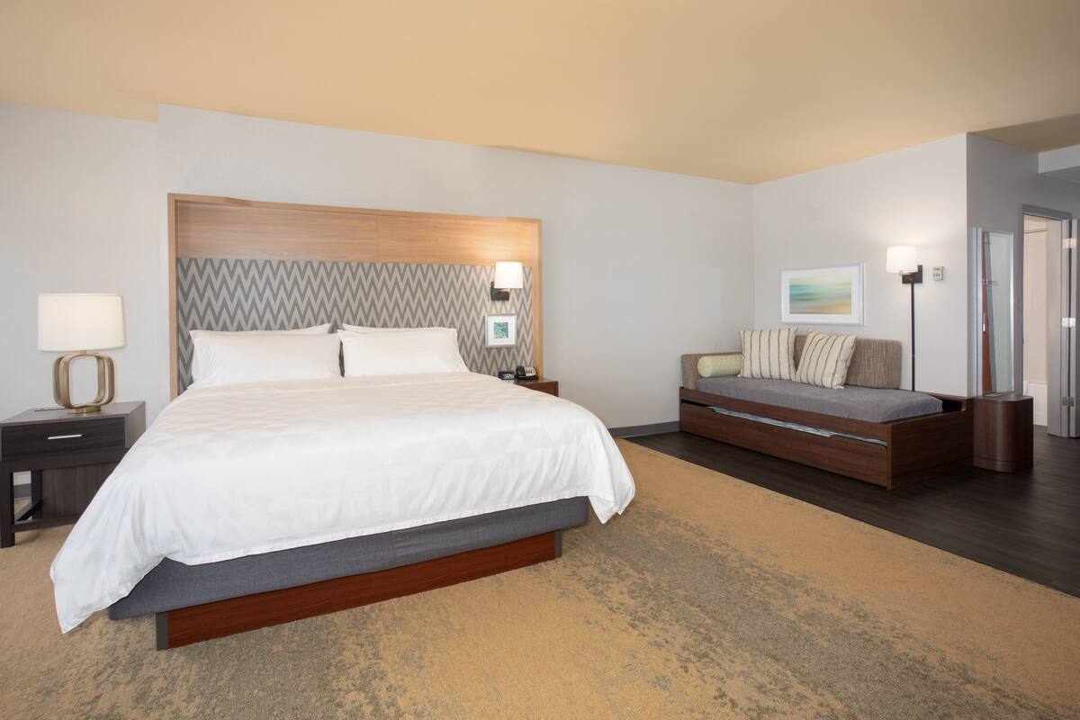 A modern hotel room with a large bed, a sofa, a bedside table with a lamp, and a wall-mounted artwork.