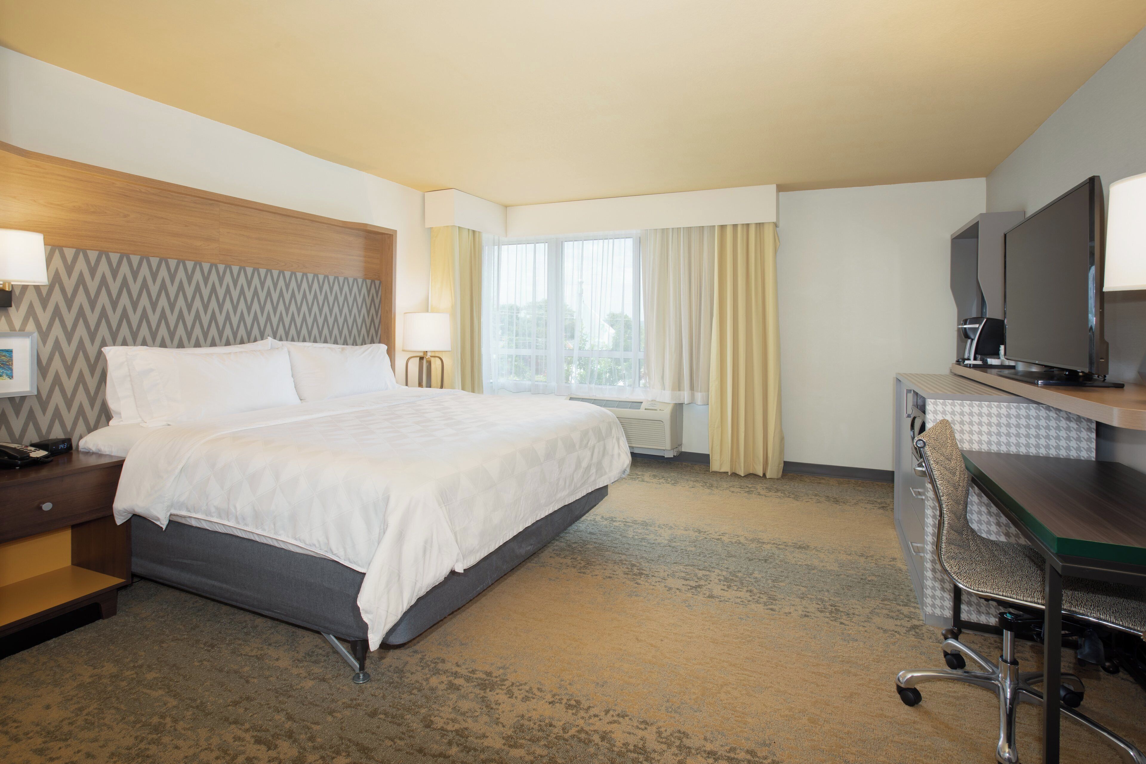 Habitación de hotel con cama, mesita de noche, lámpara, escritorio con silla, televisor y ventana con cortinas.