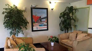 Un vestíbulo de hotel con dos sofás color beige, una mesa de centro y una planta grande en maceta. En la pared hay un cartel enmarcado de un coche y un edificio.