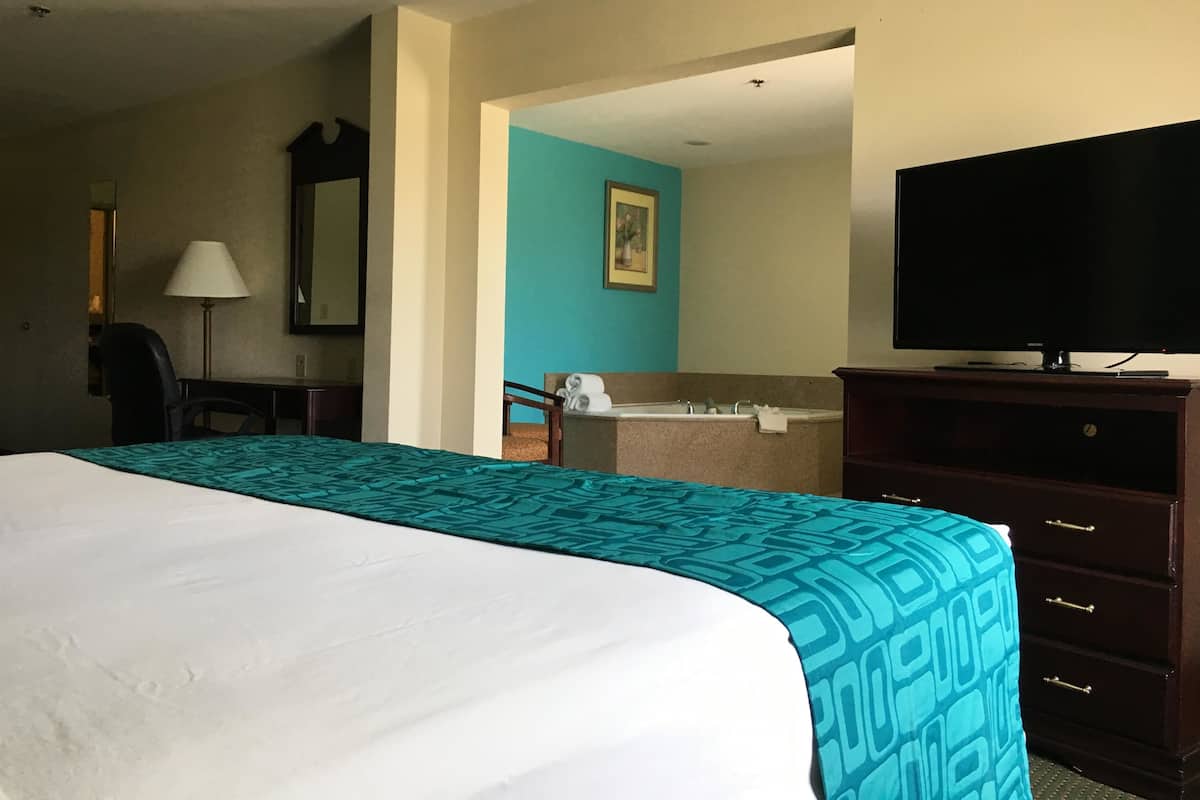 A hotel room with a large bed, a desk with a lamp, a television, a bathroom with a sink and mirror, and a painting on the wall.
