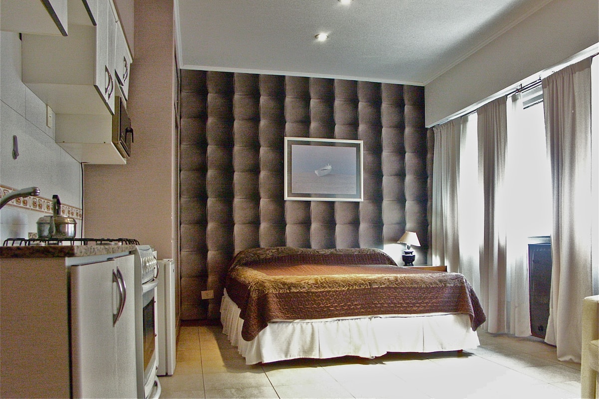 Un dormitorio con cama, mesita de noche, lámpara, ventana con cortinas, un cuadro en la pared y una kitchenette con refrigerador y estufa.