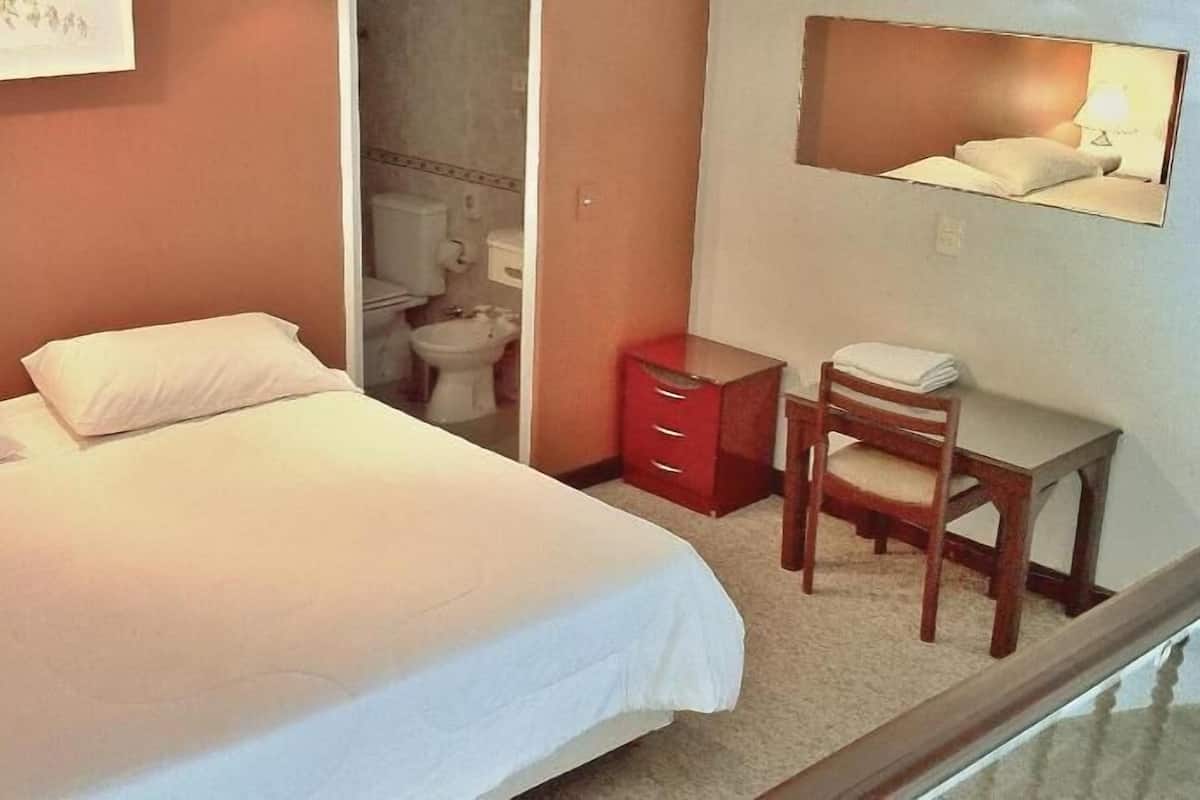 Habitación de hotel con cama, escritorio con silla, cómoda roja y baño visible a través de una puerta abierta.