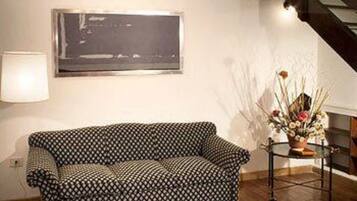 A living room with a patterned sofa, a framed picture on the wall, a lamp, and a small table with a vase of flowers.