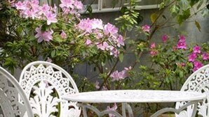 Une table et des chaises en métal blanc, placées à l’extérieur, avec des fleurs roses en arrière-plan.