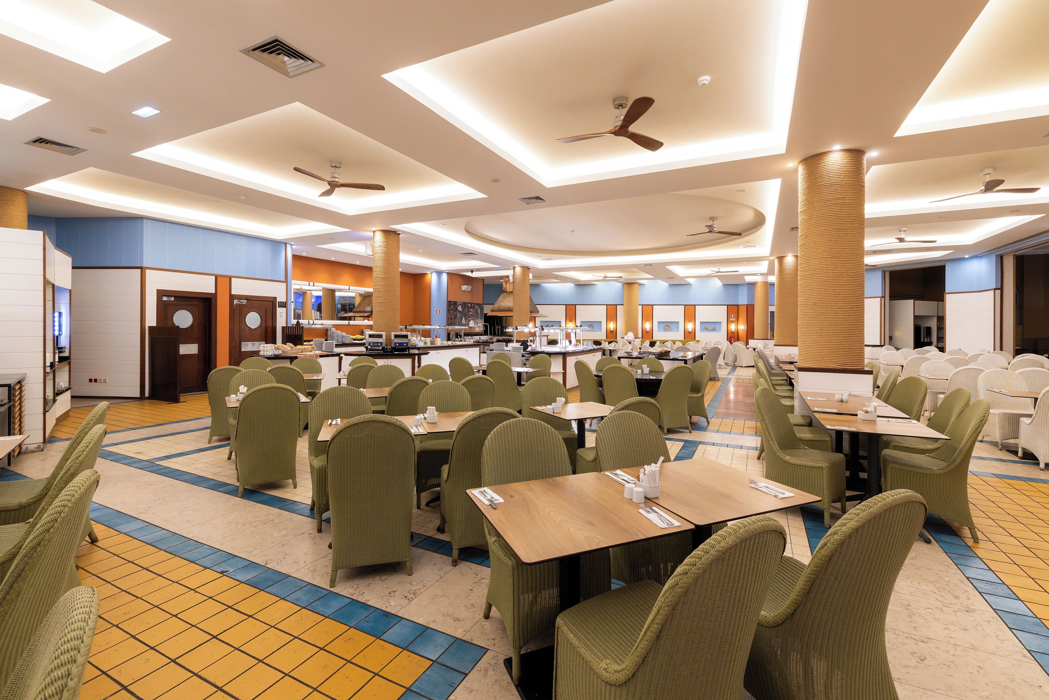 A spacious restaurant with high ceilings, multiple dining tables, and green upholstered chairs.