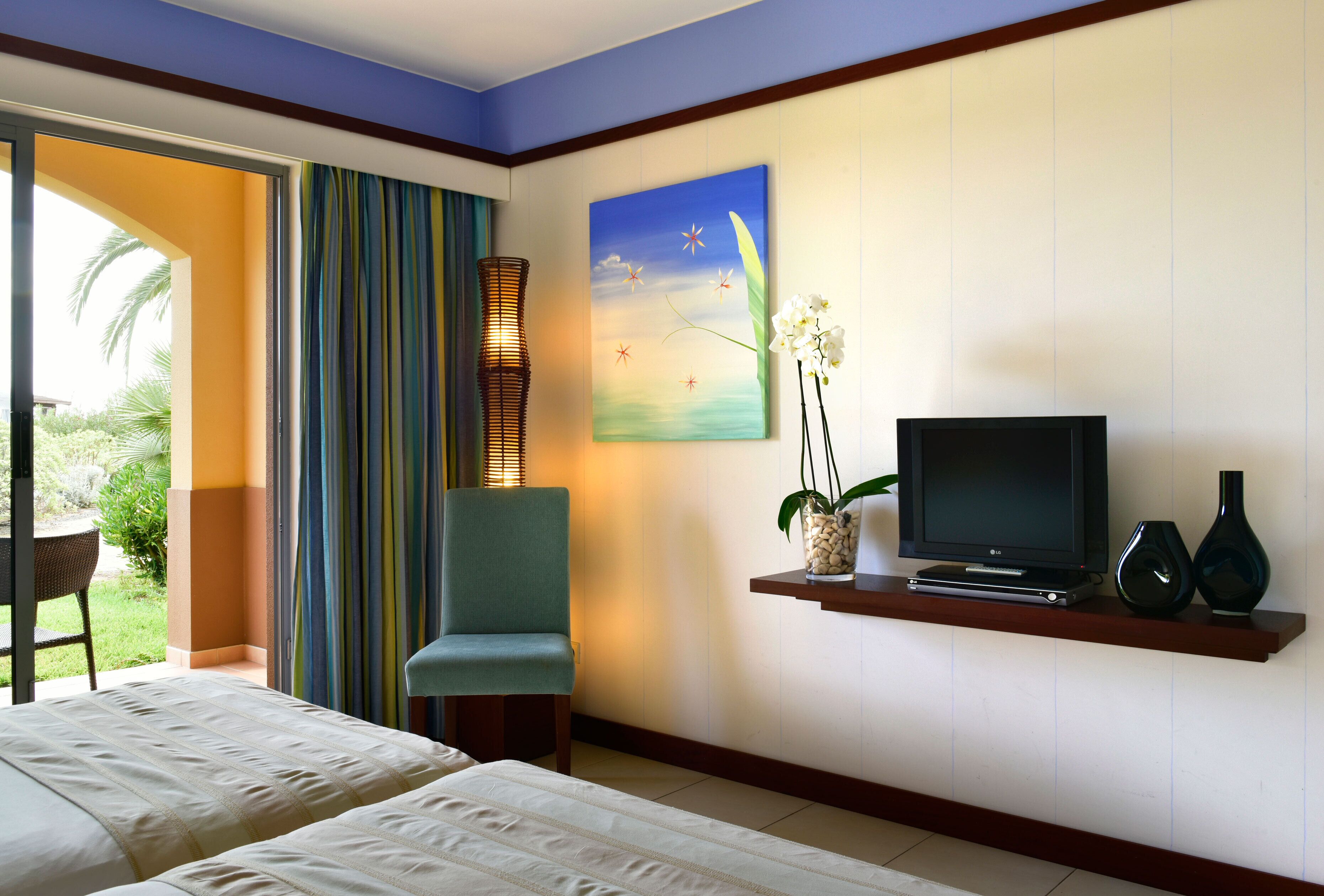 A hotel room with a bed, a television, a chair, a painting, and a view of the outdoors.