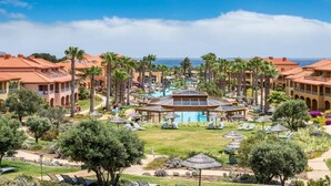 Un complejo turístico con piscina central, palmeras y edificios de estilo mediterráneo.