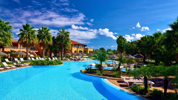 Zona de piscina en un resort con sillones de descanso, palmeras y un edificio grande con techo rojo.