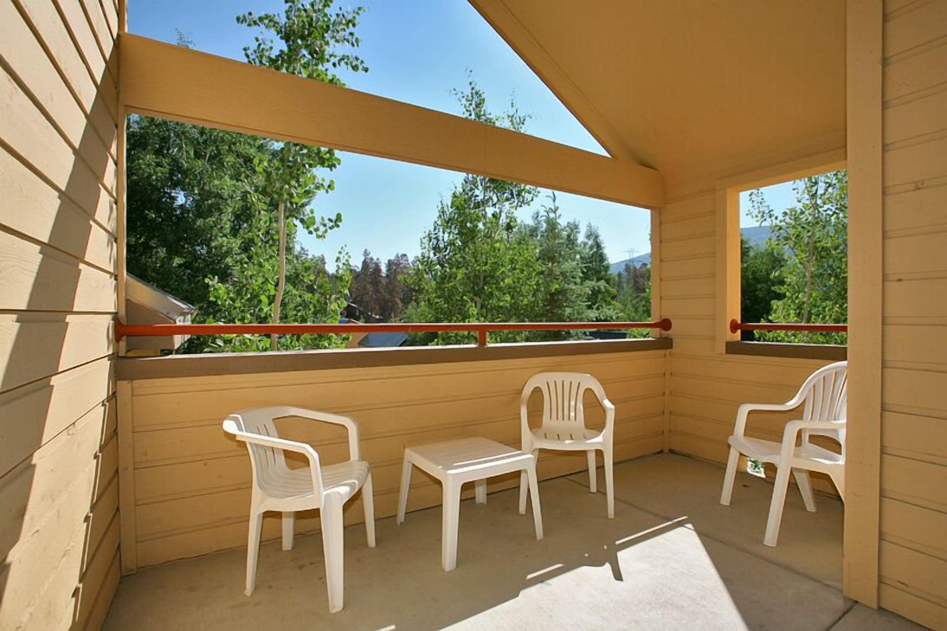 A covered balcony with three white plastic chairs and a small table, overlooking a green landscape with trees and a clear sky.