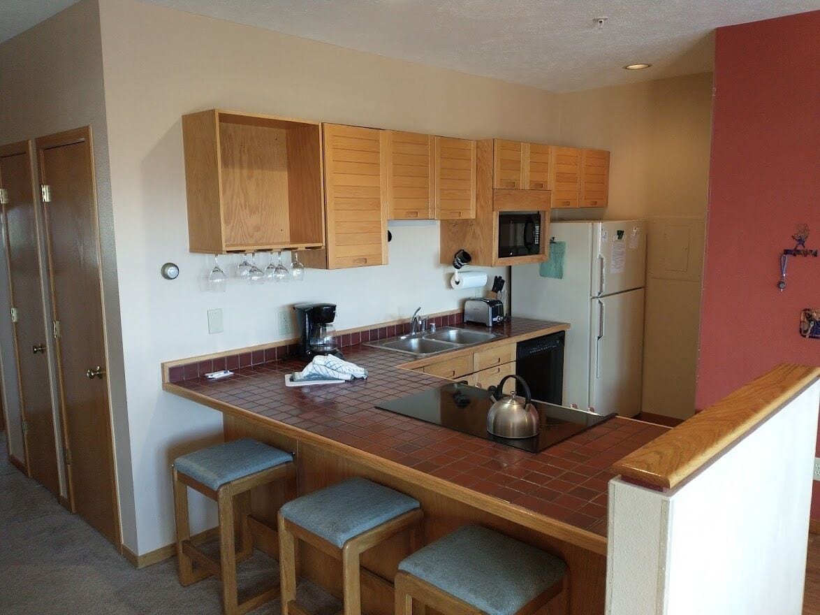 A compact kitchen with wooden cabinets, a microwave, a refrigerator, and a small island with stools.