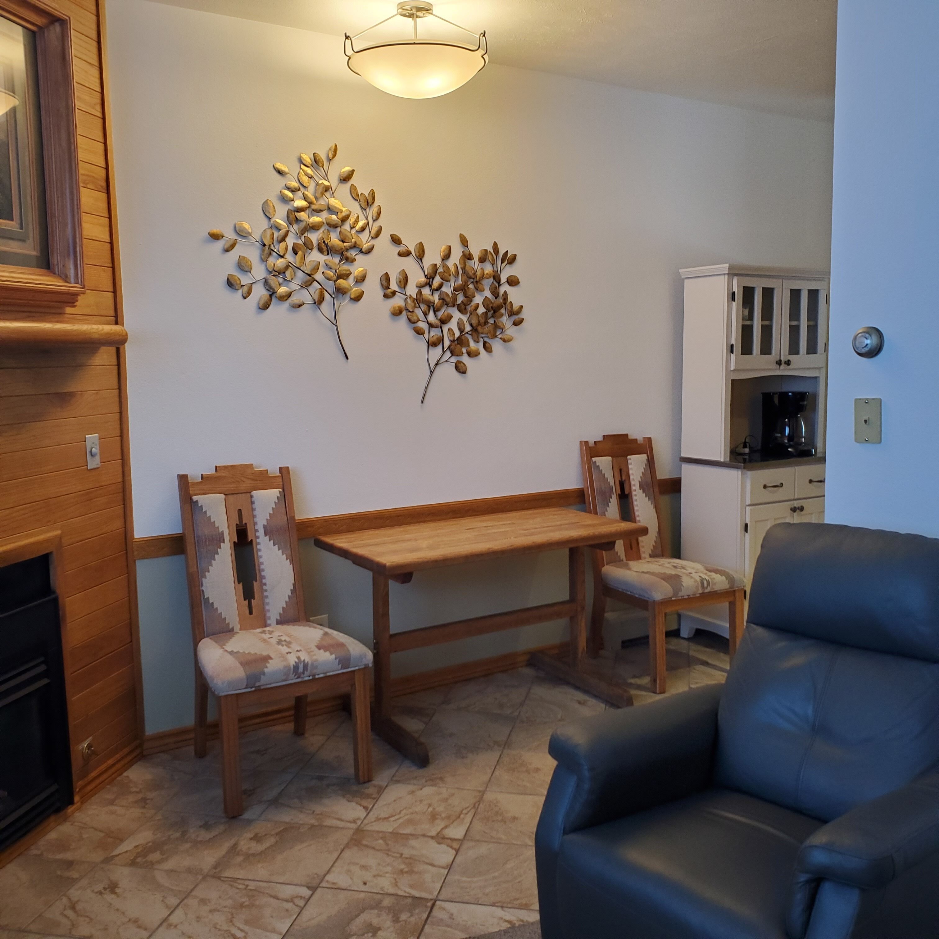 A dining area with a wooden table and chairs, a decorative wall piece, and a coffee machine in the background.