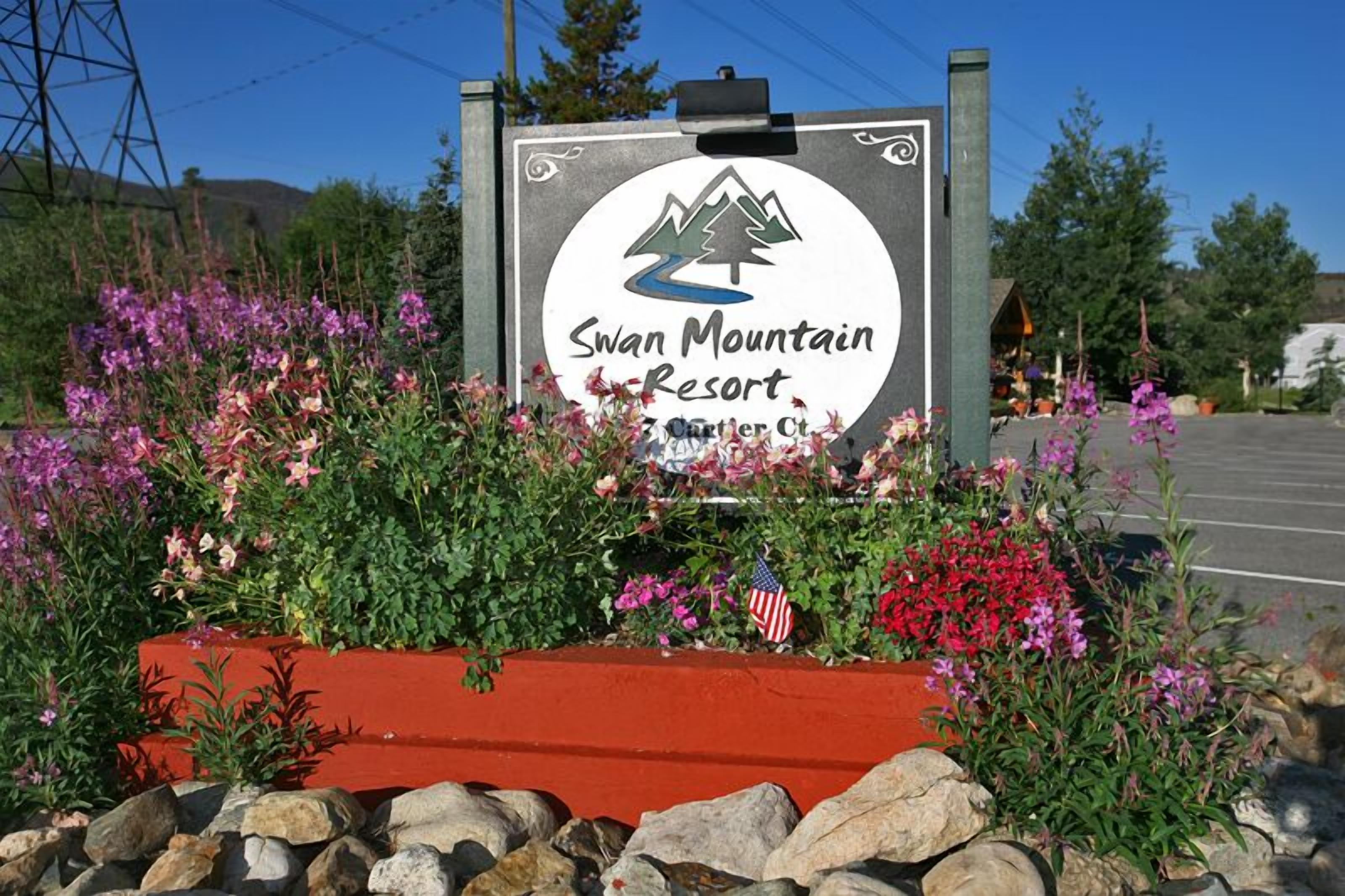 Señal para Swan Mountain Resort, con un logotipo de montaña, rodeada de flores coloridas y rocas.