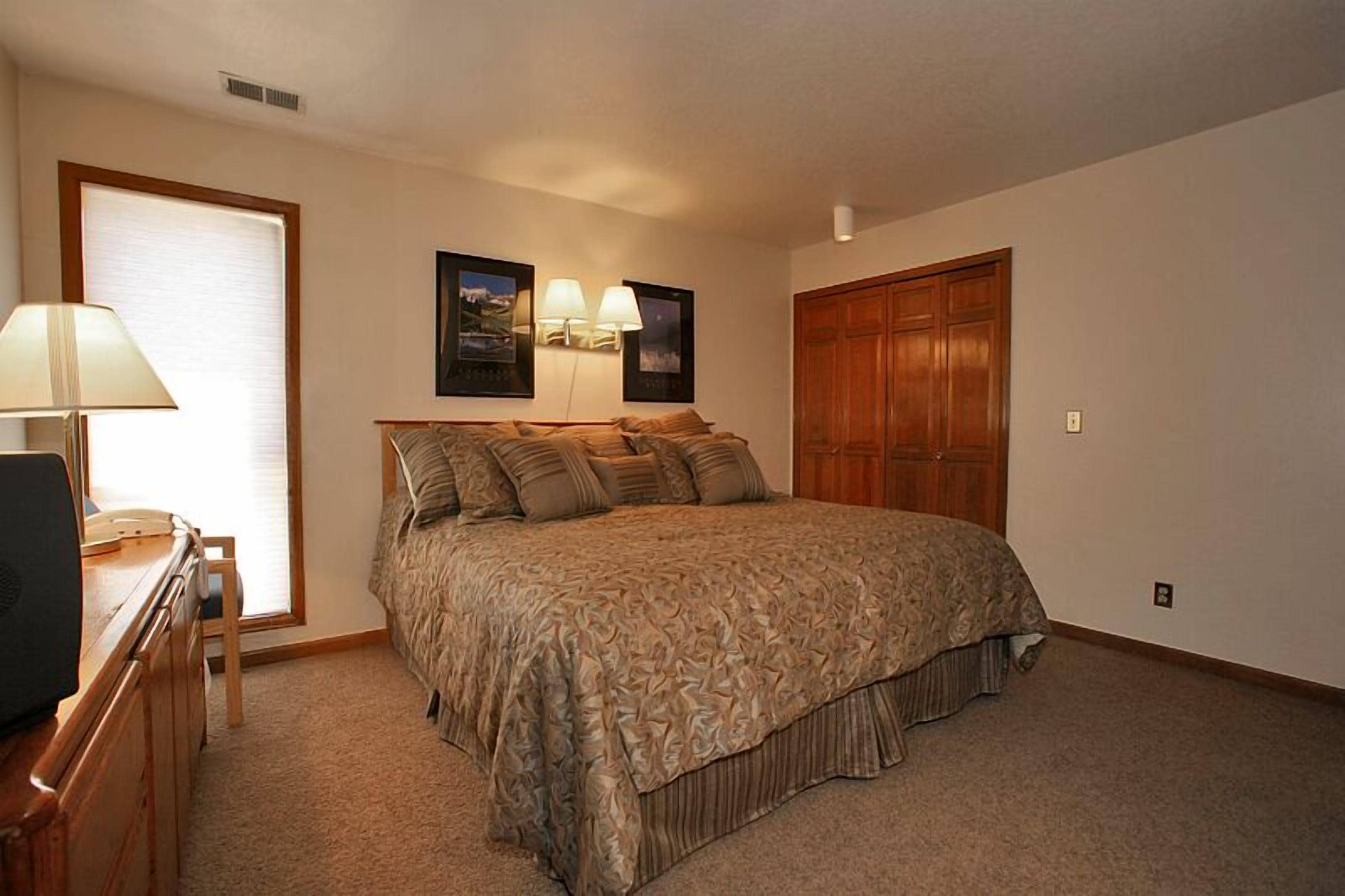 A bedroom with a large bed, a desk with a lamp, two wall-mounted lamps, and a wooden wardrobe.