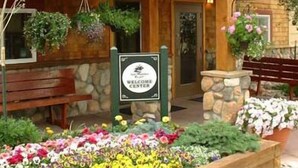 A wooden building with a 'Welcome Center' sign, stone walls, benches, and flower beds. - Swan Mountain Resort (Dillon)