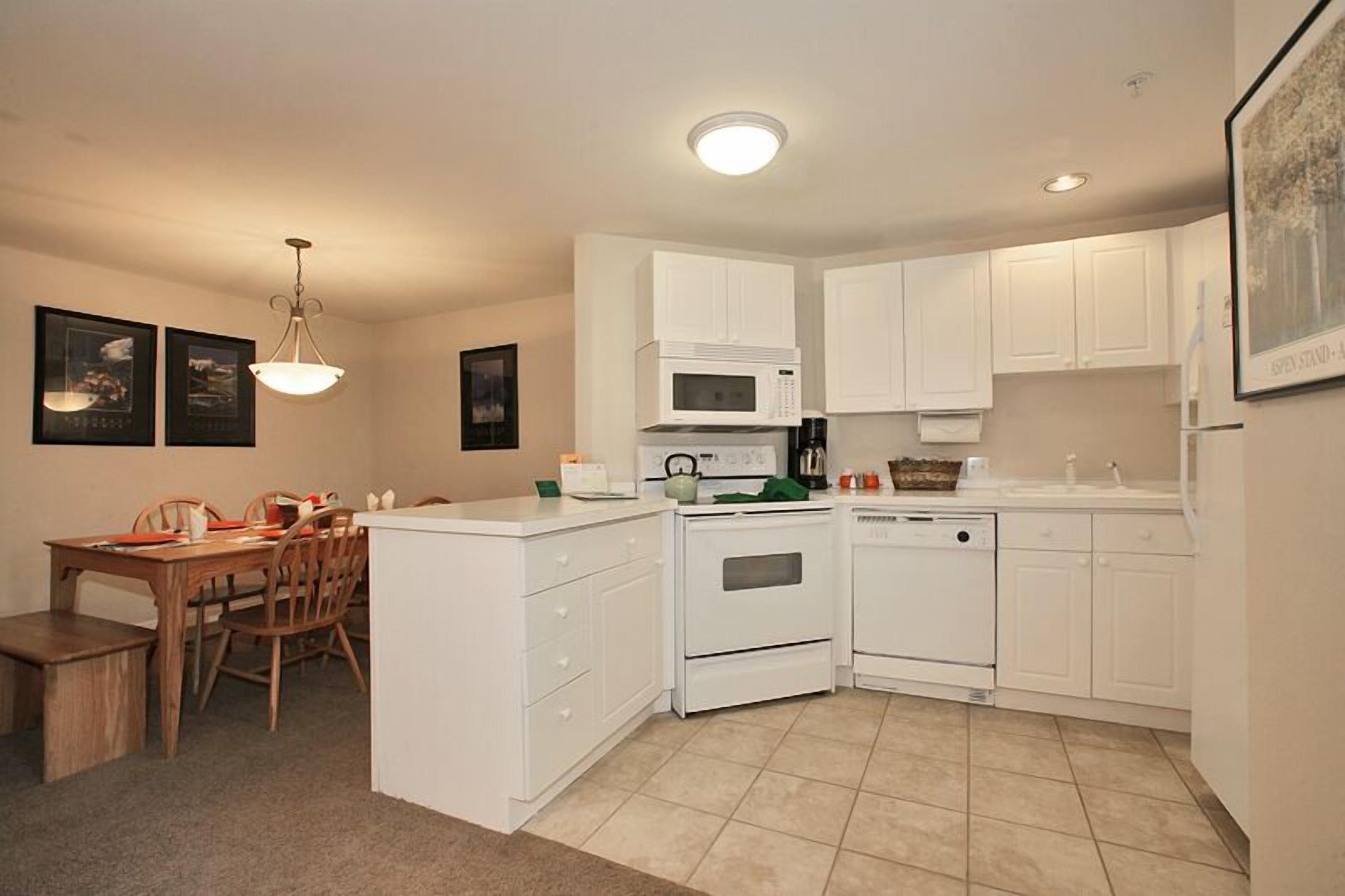 A compact kitchen with white cabinets, a microwave, an oven, and a dishwasher. There is a dining table with chairs and a hanging light fixture.