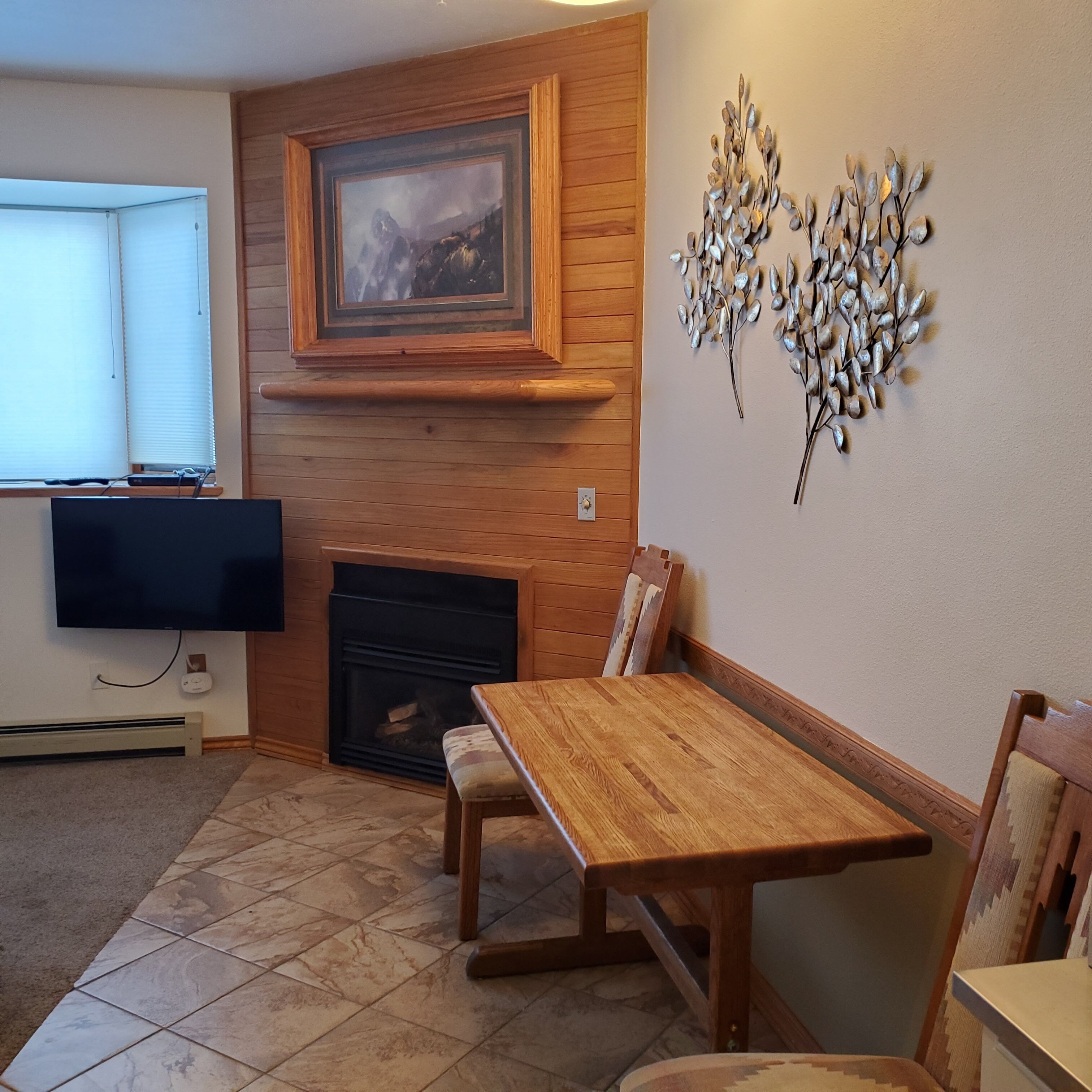 A cozy living room with a fireplace, wooden paneling, a dining table, and a TV mounted on the wall.