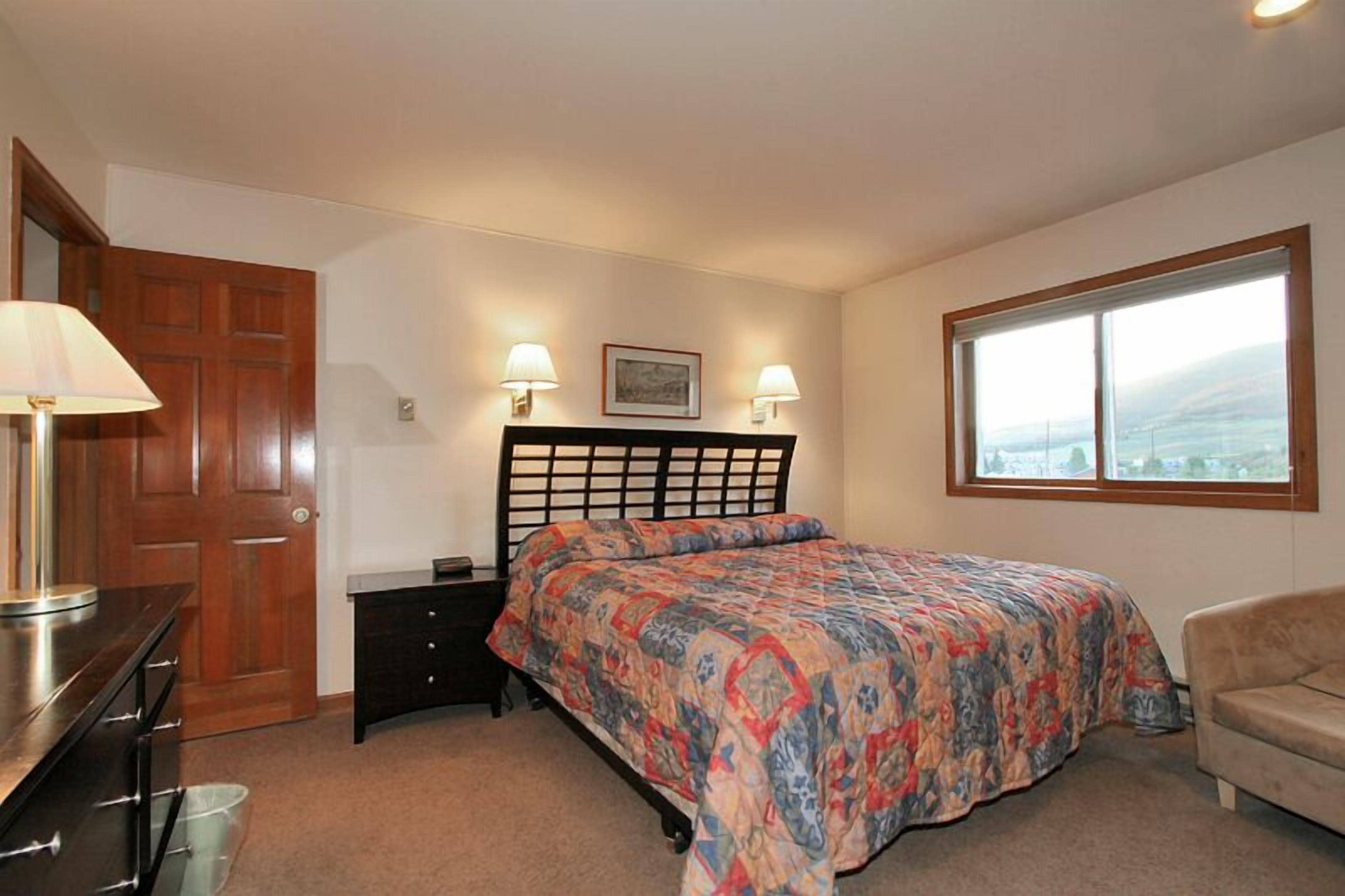 A bedroom with a bed, a wooden door, a dresser, a nightstand, a lamp, a window, and a picture on the wall.