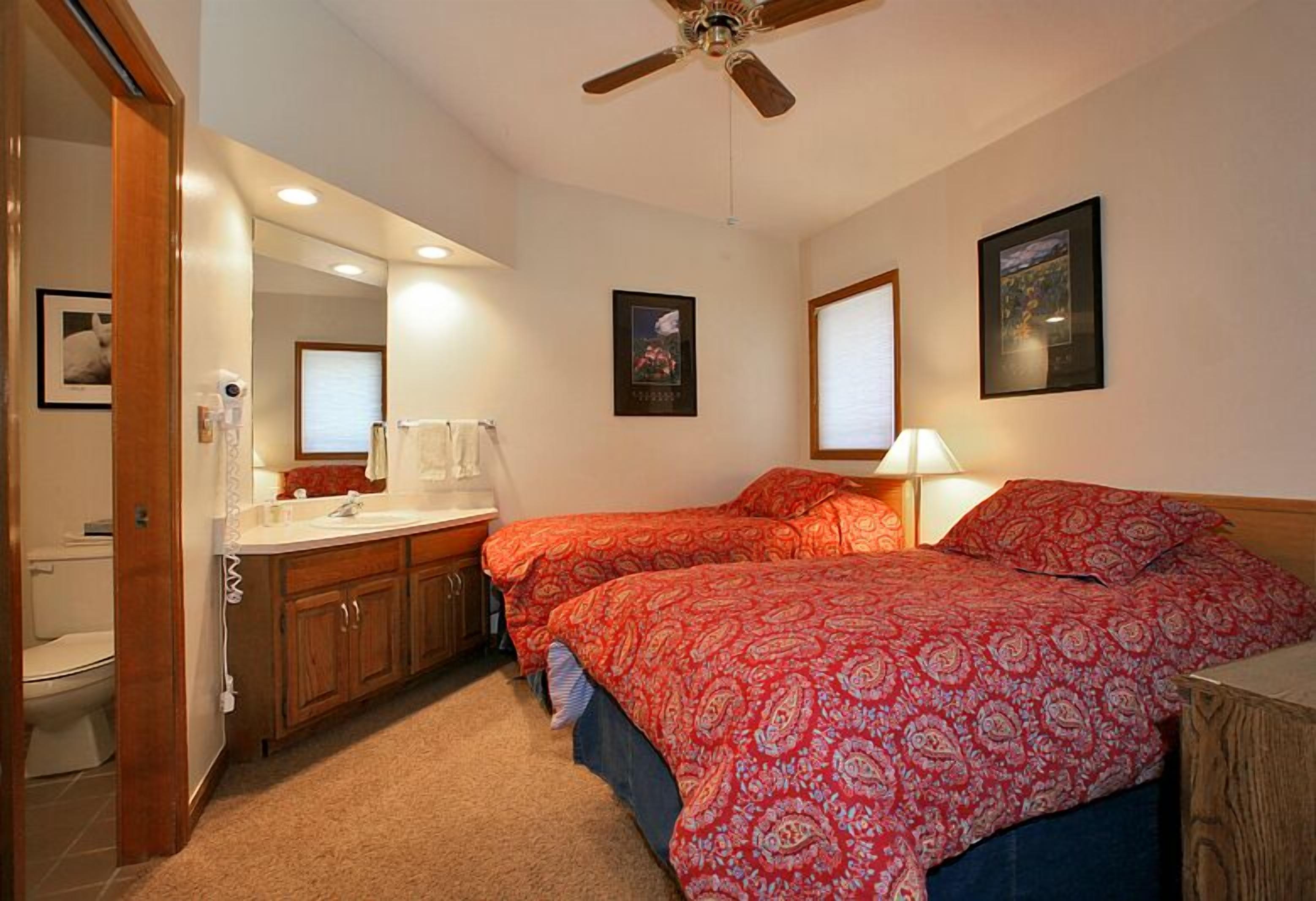 A bedroom with two beds, a ceiling fan, a bathroom with a toilet, and a sink.