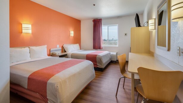 Habitación de hotel con dos camas, un escritorio y una silla. Cuenta con una ventana con cortinas, un espejo y luces en la pared.