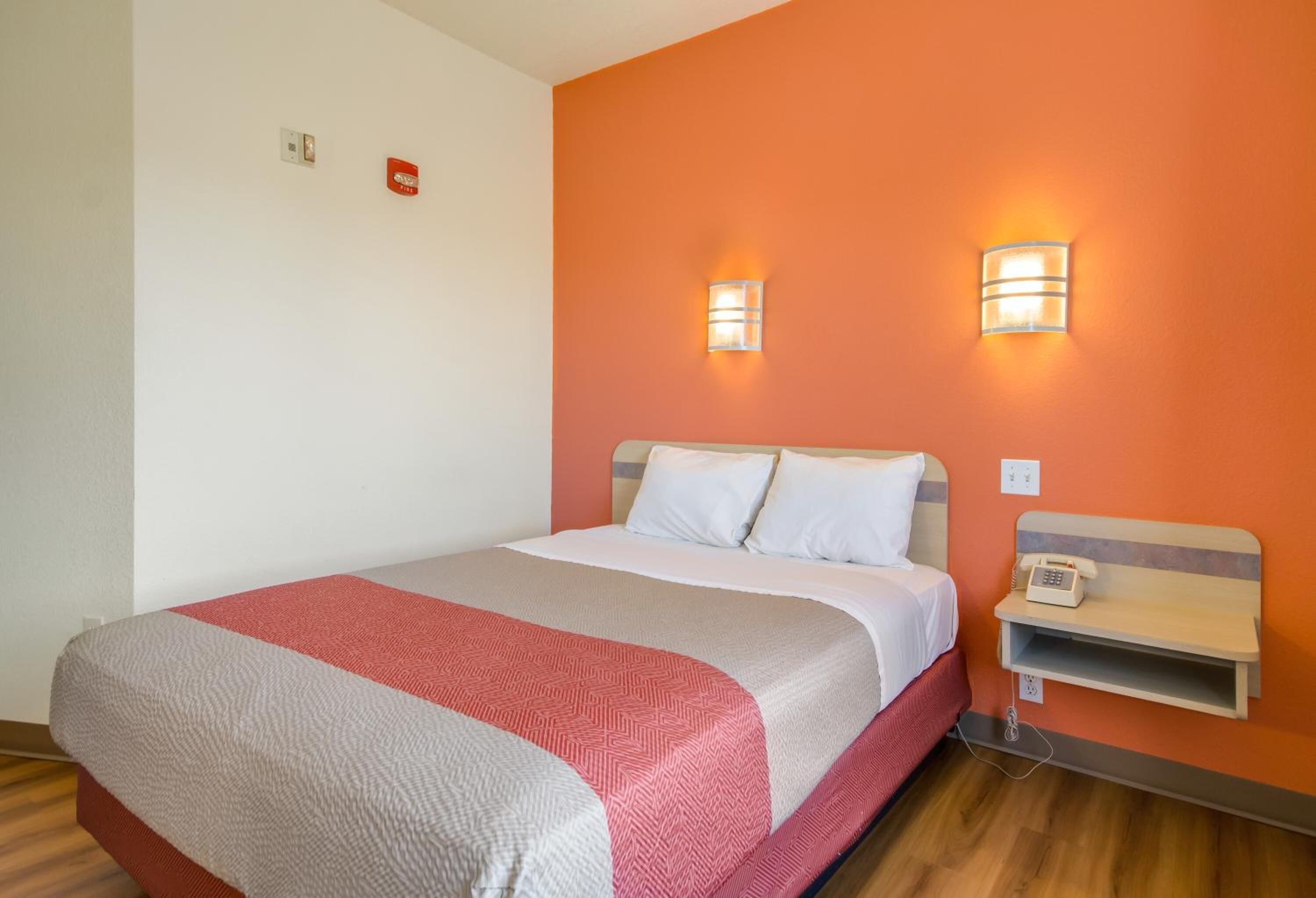 A hotel room with a bed, two wall-mounted light fixtures, a bedside telephone, and a small shelf.