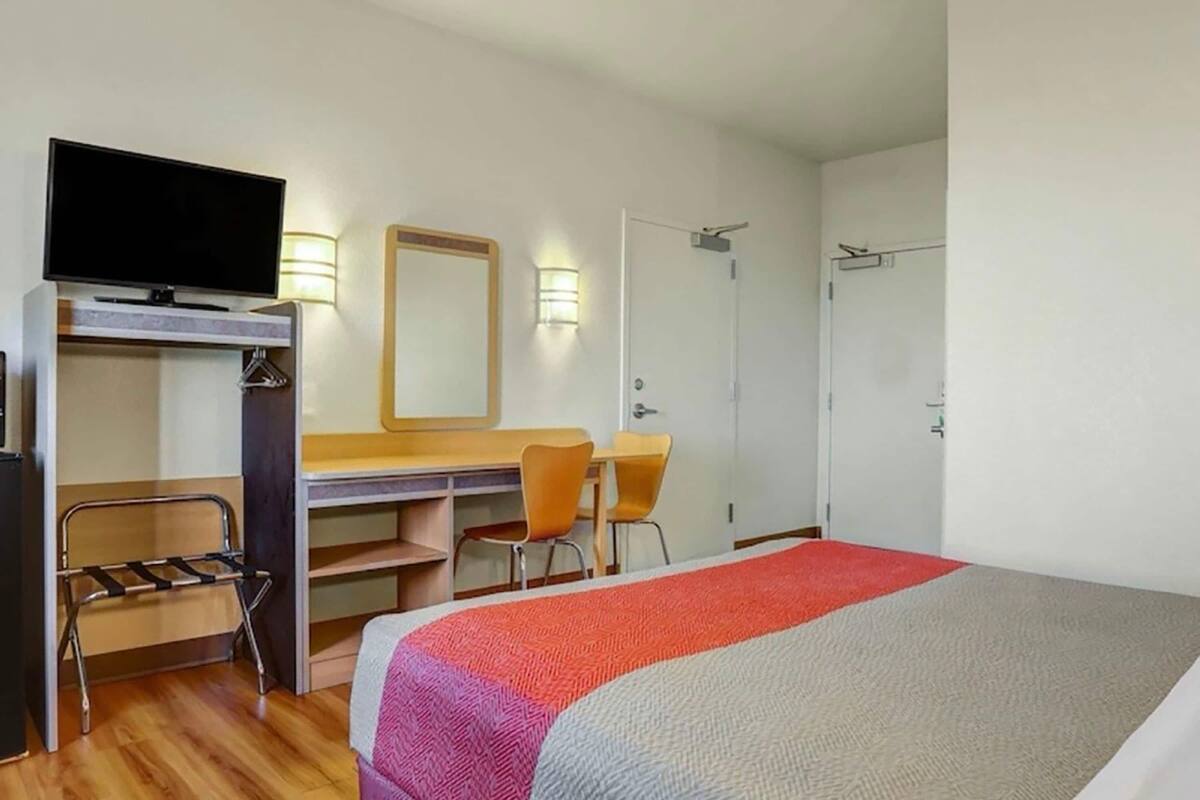 A hotel room with a bed, a desk with a chair, a television, a mirror, and a wardrobe.