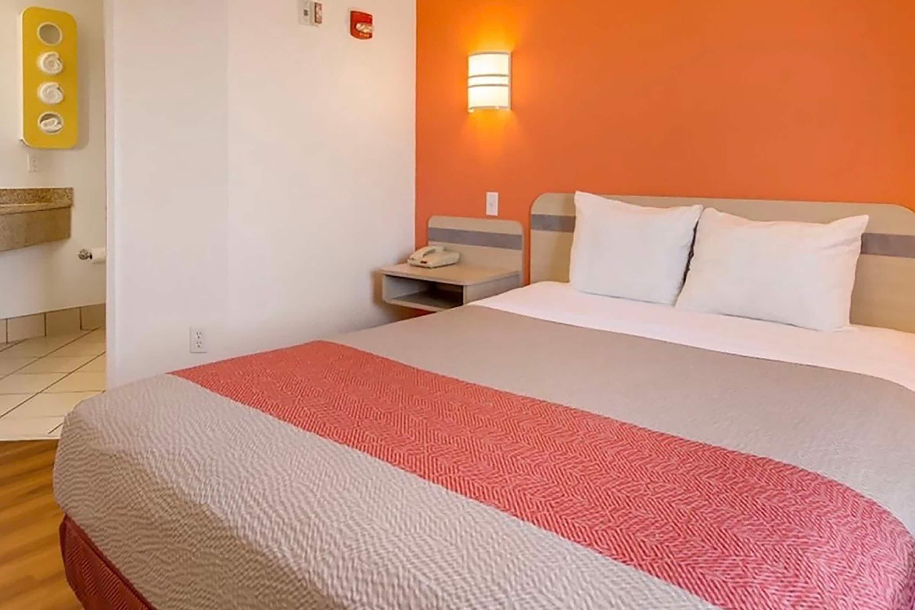 A hotel room with a bed, a nightstand, a wall-mounted light, and a bathroom with a towel rack.