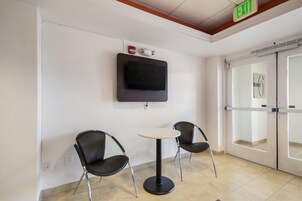 Une salle d’attente avec deux chaises noires, une petite table ronde, un téléviseur à écran plat fixé au mur et un panneau indiquant la sortie.