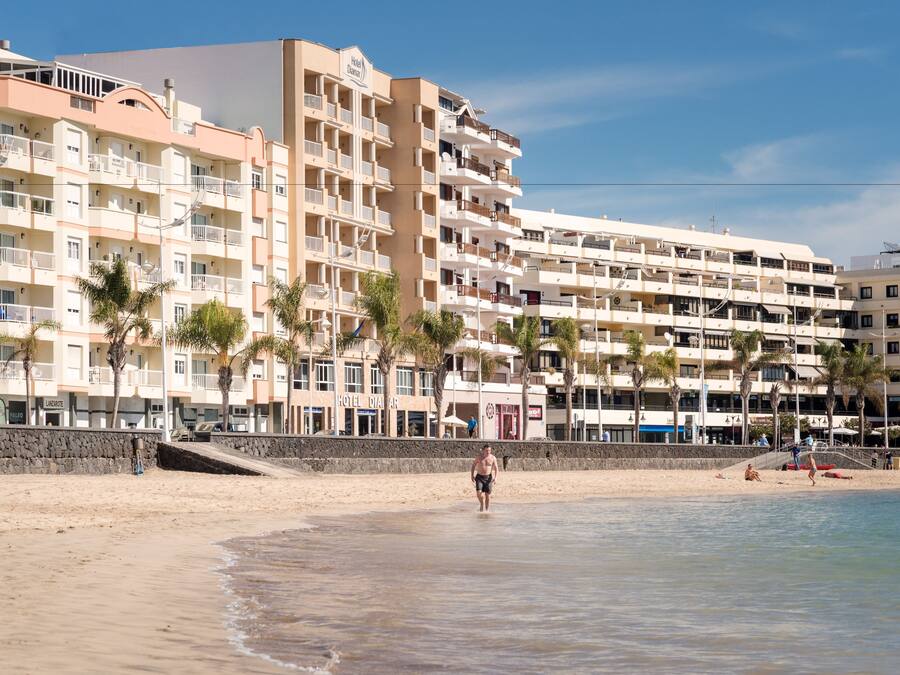 Un hotel frente a la playa con varios balcones, una playa de arena y palmeras.