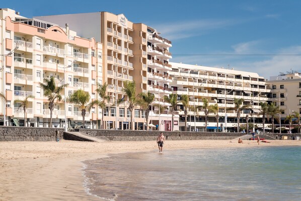 Un hotel frente a la playa con varios balcones, una playa de arena y palmeras.