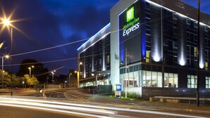 Un bâtiment de l’hôtel Holiday Inn Express doté d’une façade moderne en verre, situé à un carrefour où l’on peut voir des feux de signalisation et des lampadaires.