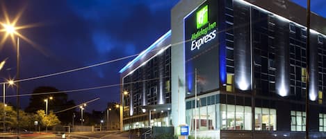 Edificio de Holiday Inn Express con fachada moderna de vidrio, situado en una intersección con semáforos y farolas.