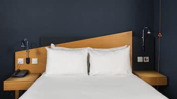 Una habitación de hotel con cabecera de madera, ropa de cama blanca, una mesita de noche con lámpara, teléfono y toma de corriente.