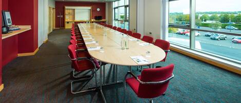 Un salón de conferencias con una mesa larga, sillas rojas y ventanas grandes que dan a un estacionamiento.