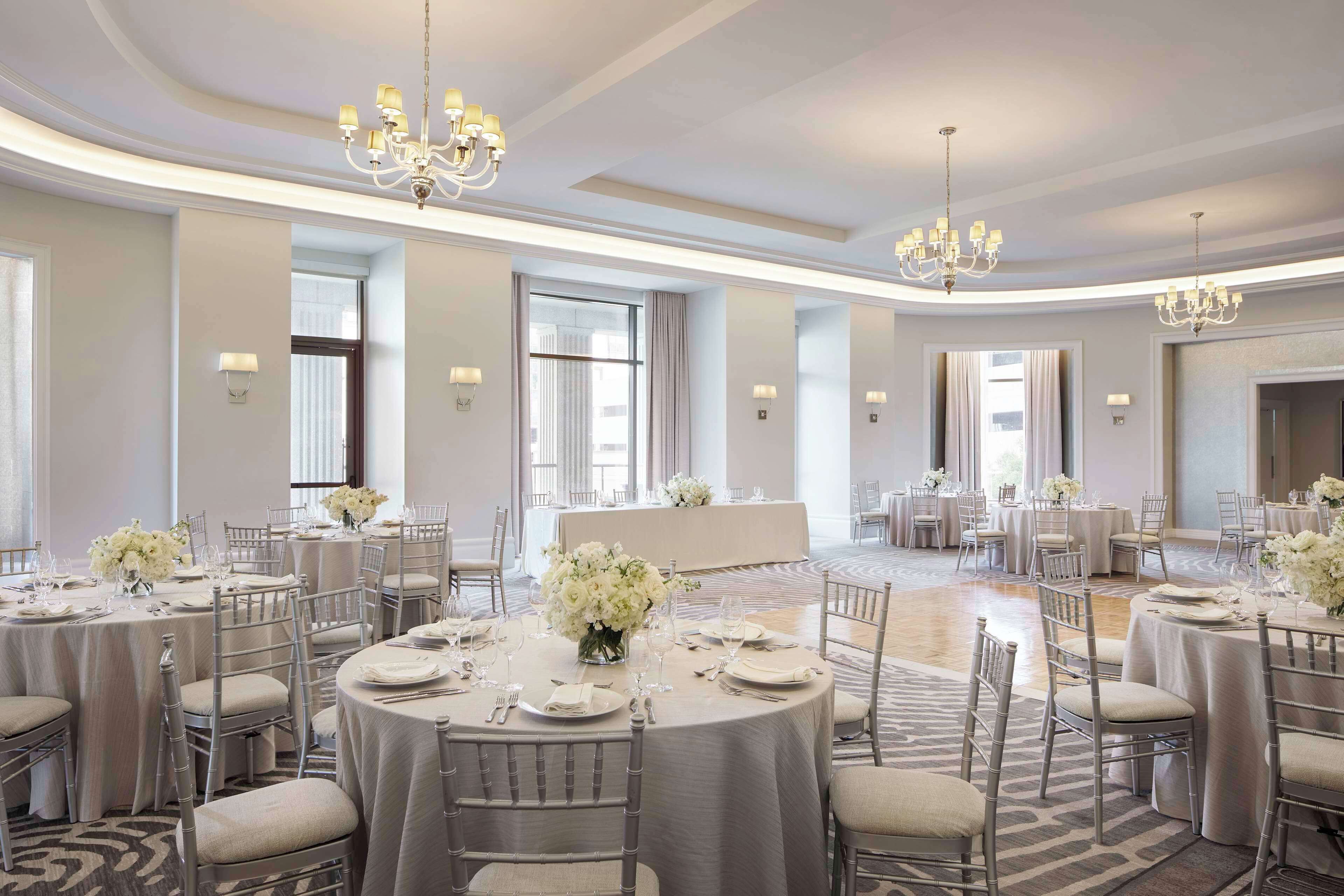 Une salle de banquet spacieuse, dotée de tables rondes, d’élégants lustres et de compositions florales blanches.