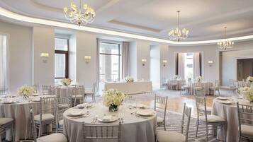 Une salle de banquet spacieuse, dotée de tables rondes, d’élégants lustres et de compositions florales blanches.