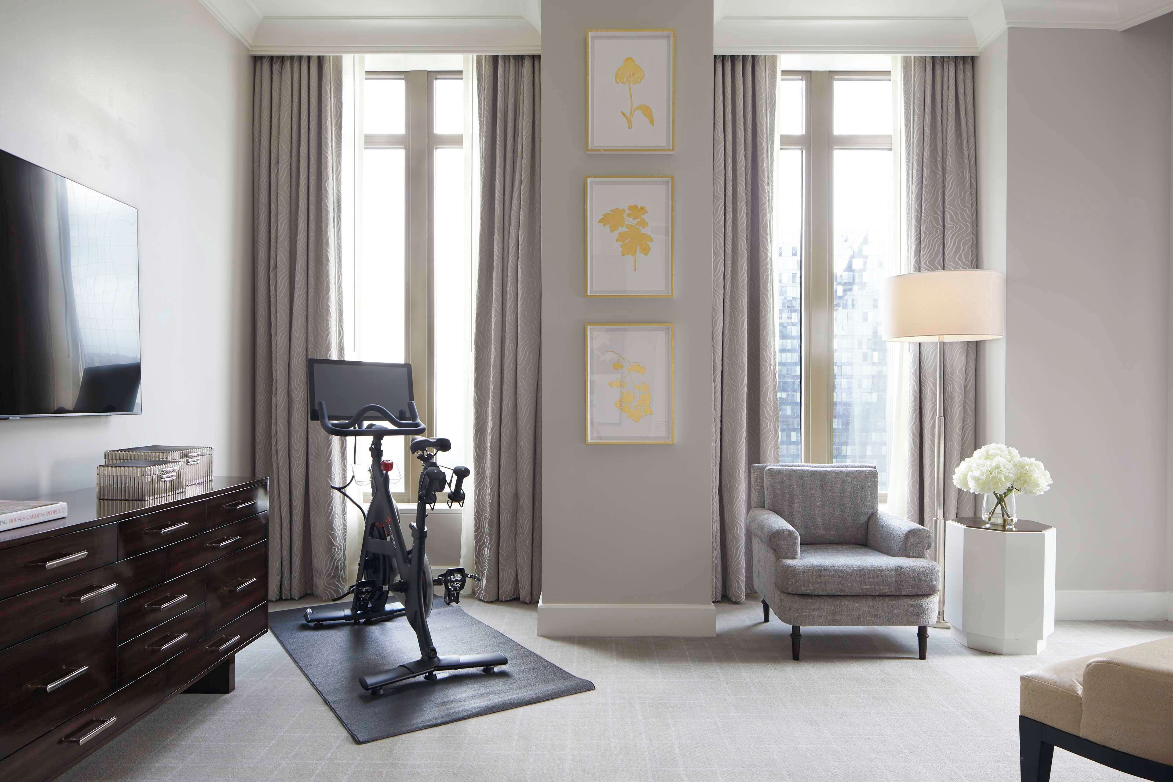 Una habitación de hotel moderna con una televisión de pantalla plana grande, equipo de ejercicio, un sillón gris y una mesita de noche con una lámpara y flores.
