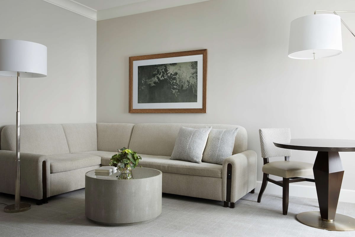 A modern living room with a beige sofa, a round coffee table with a vase, a framed picture on the wall, and a round table with a chair.