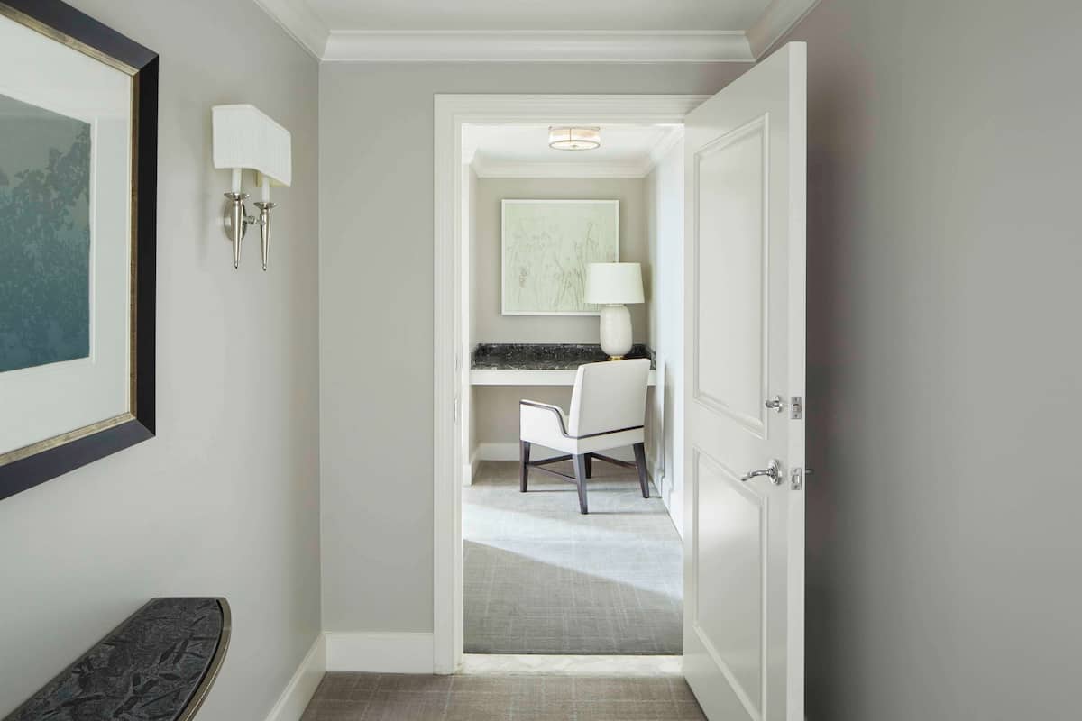 A hallway leading to a bathroom with a white chair, a framed artwork on the wall, and a light fixture.