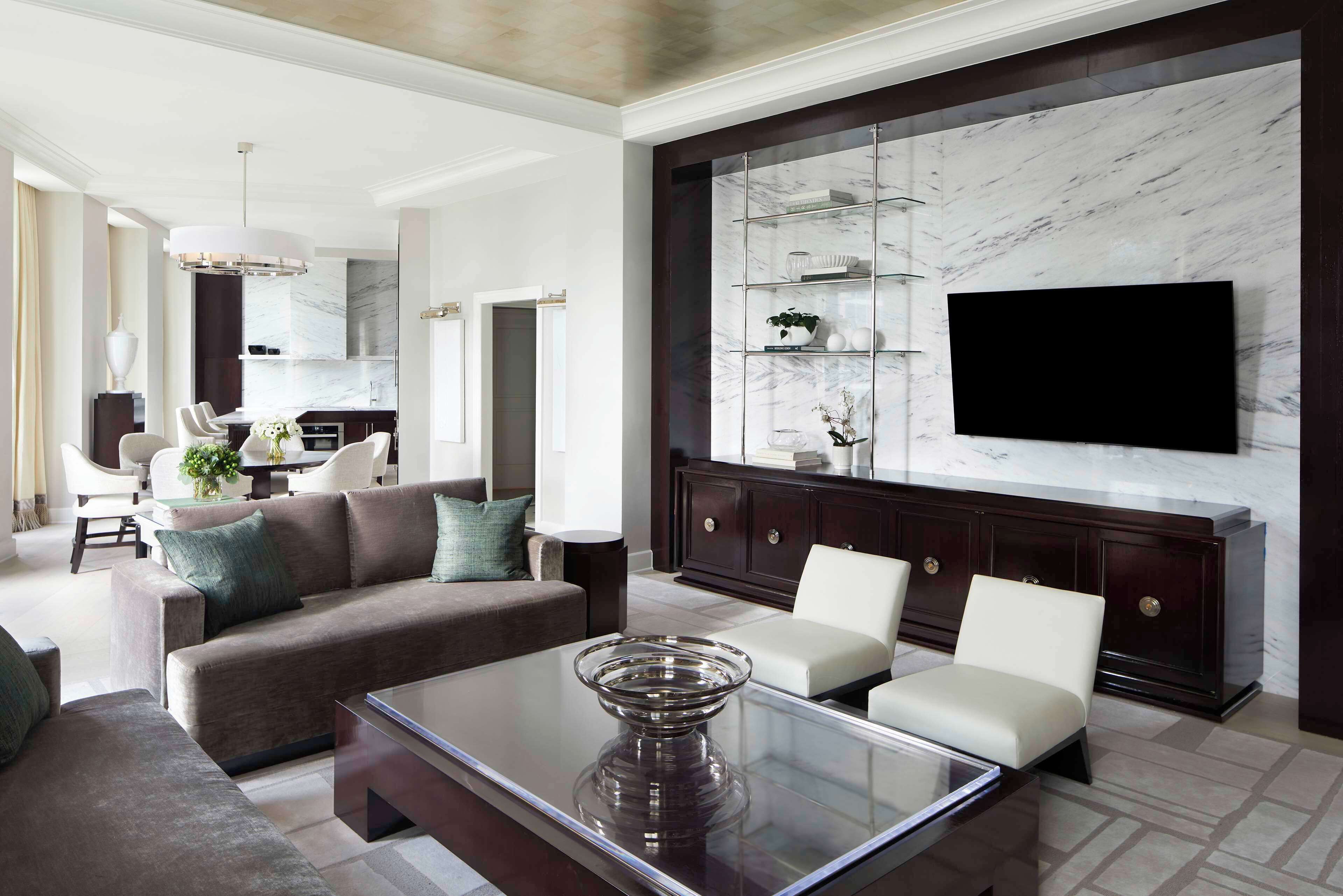 A modern living room with a glass coffee table, a dark wood cabinet with shelves, and a flat-screen TV mounted on the wall.