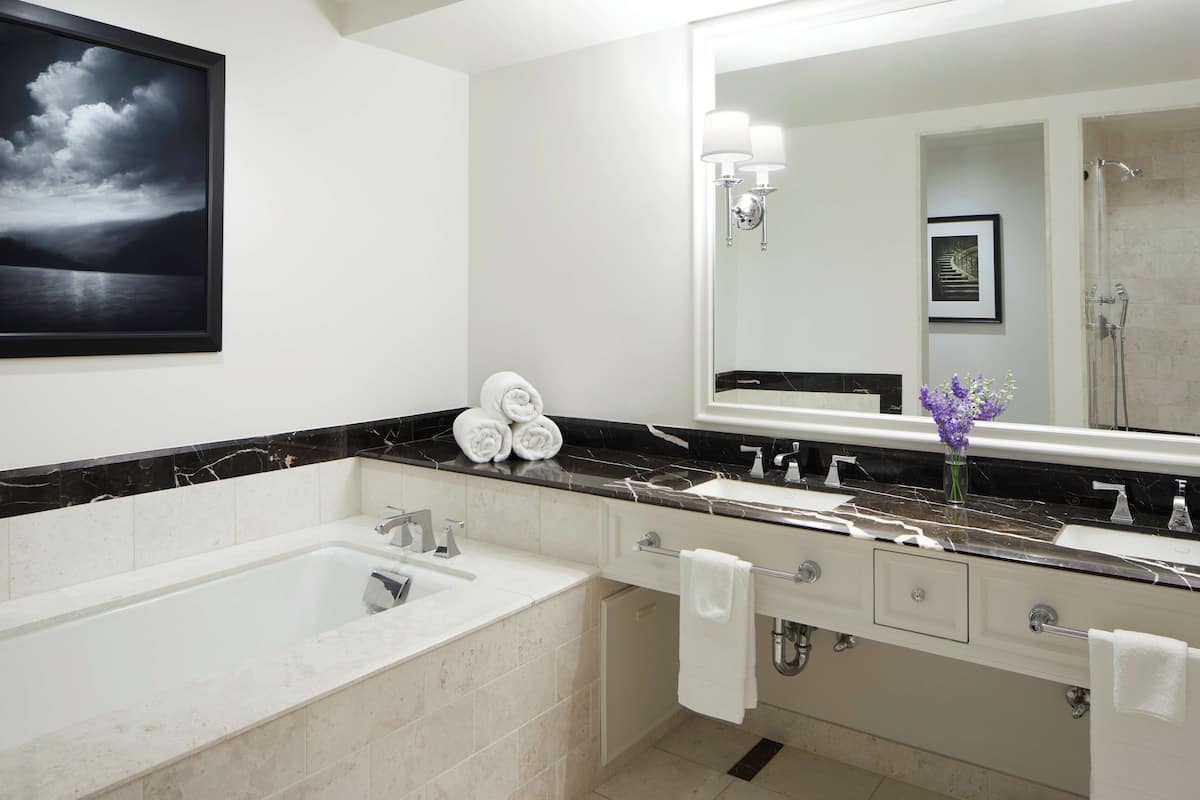 A modern bathroom with a large bathtub, two sinks, and a large mirror. There are towels and a vase with flowers on the counter.