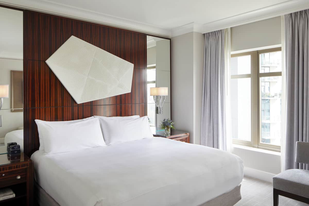 A hotel room with a large bed, a wooden headboard, a nightstand with a lamp, a mirror, and a window with curtains.