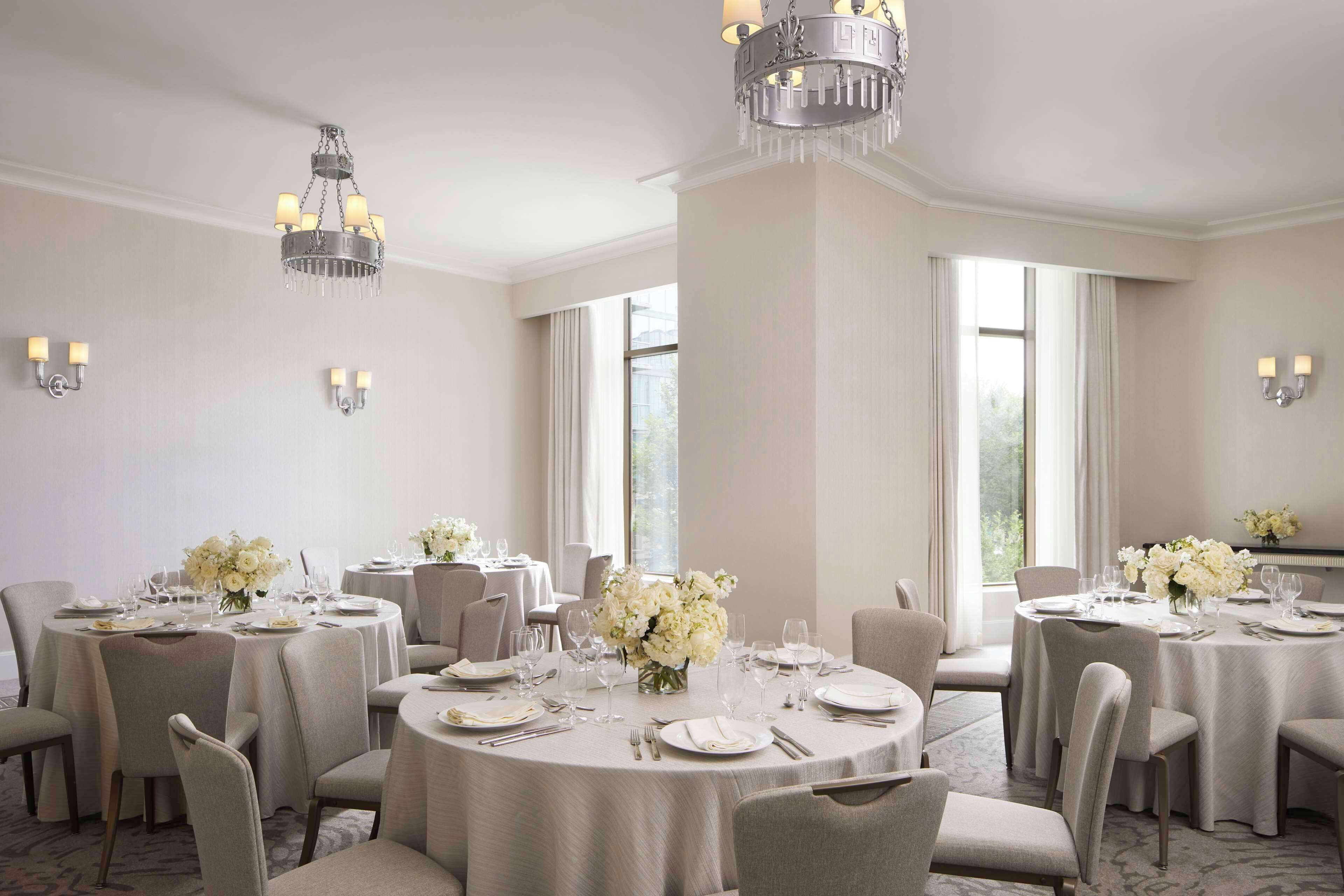 En hotels restaurant med runde borde dækket til servering, elegante lysekroner og store vinduer der lukker naturligt lys ind.