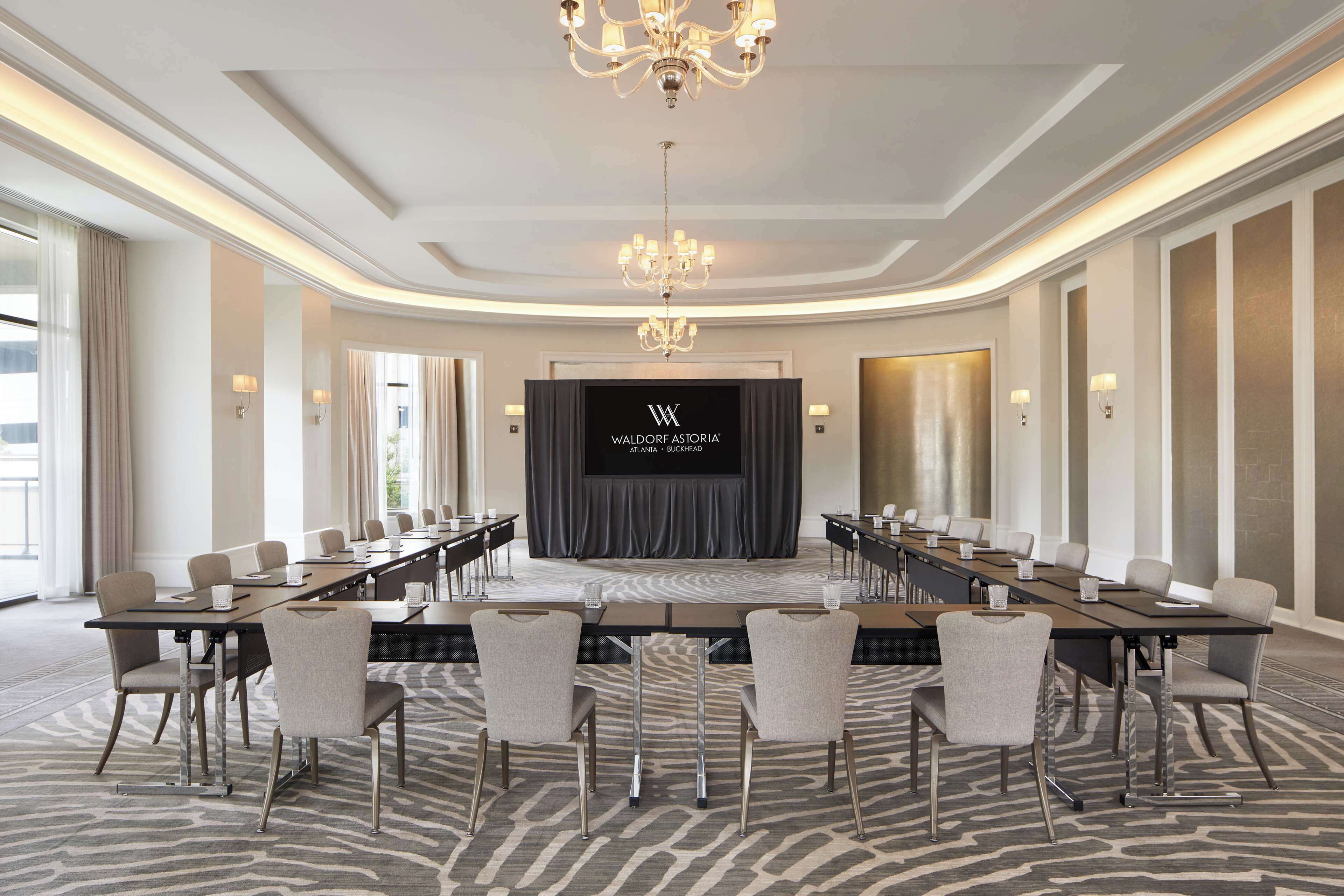 Un salón de conferencias con una larga mesa, sillas y un escenario con una pantalla que muestra 'Waldorf Astoria Atlanta'.
