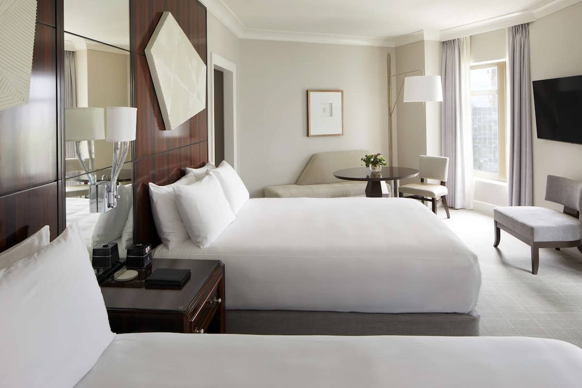 A hotel room with a large bed, a desk with a lamp, a chair, a small table with a plant, and a TV on the wall.