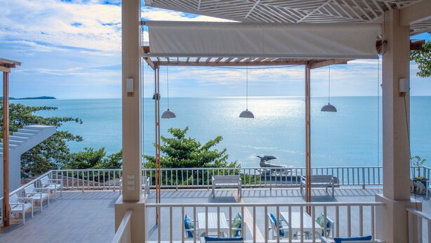 Un espacio al aire libre techado con vistas al mar, que cuenta con mobiliario blanco y lámparas colgantes.