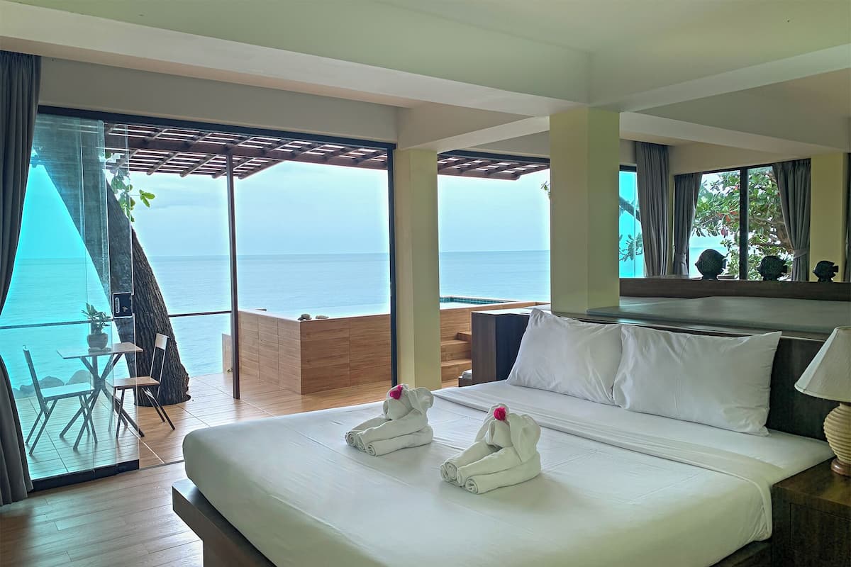 Habitación de hotel con una cama grande, vista al mar, terraza de madera y balcón con silla y mesa.