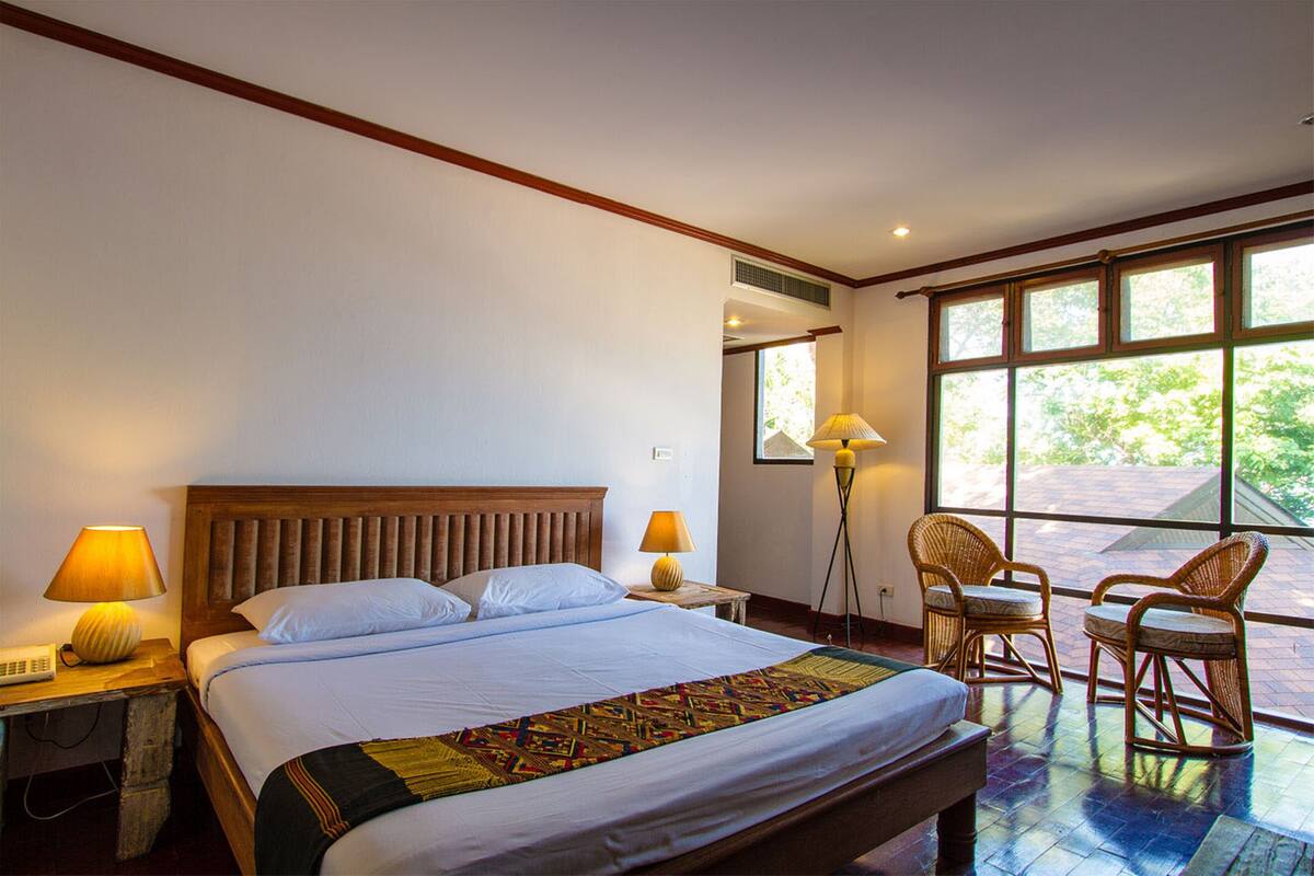Una habitación de hotel con una cama grande, dos lámparas de noche, un cabecero de madera, un ventanal con vista a la vegetación y dos sillas de mimbre.