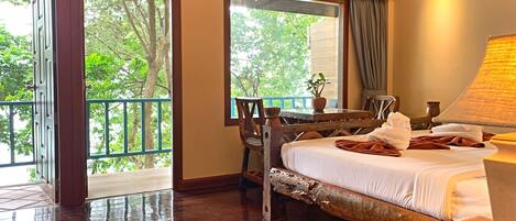 A spacious room with a large bed, a wooden desk, and a view of greenery through the windows.