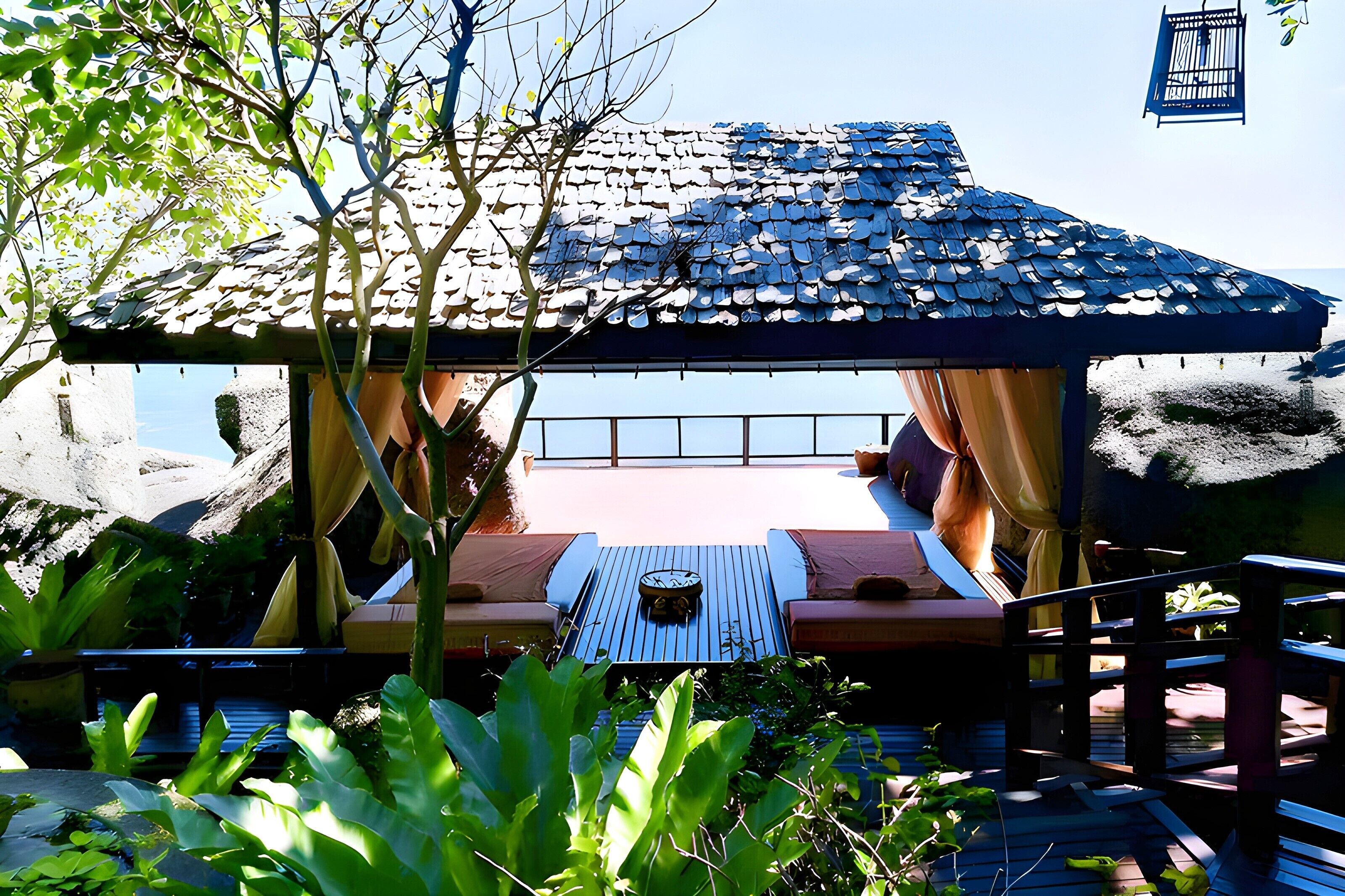 A covered outdoor seating area with a table and chairs, surrounded by plants and a view of the sea.