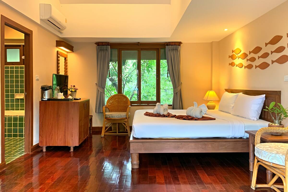 Habitación de hotel con suelo de madera, cama con ropa blanca, una silla de mimbre, un escritorio con espejo y una ventana con cortinas.